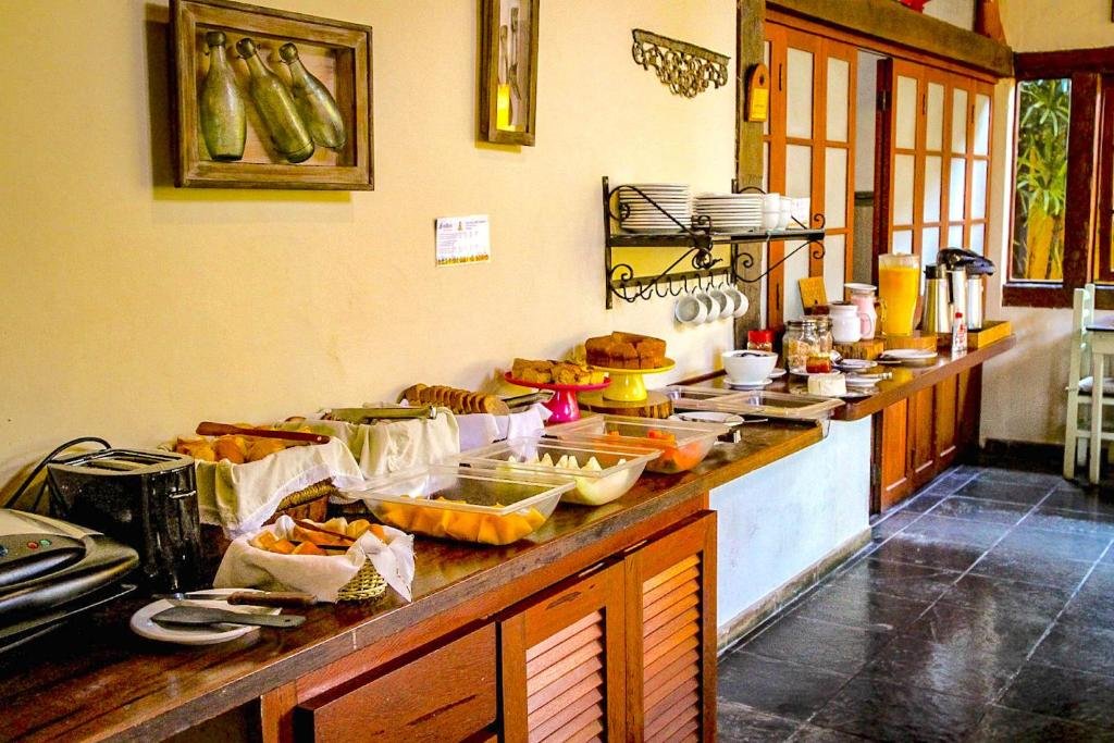 Área com mesa de café da manhã na VELINN Caravela Pousada. Há uma grande mesa de café da manhã, que conta com muitas frutas, pães, bolos, sucos, iogurte, leite e café, além de torradeira e sanduicheira.