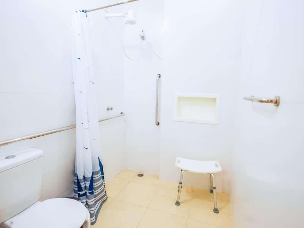 Pousada VELINN Face Norte, foto que mostra o banheiro com um vaso sanitário rebaixado à esquerda, perto de barras de apoio. Também há barras de apoio no box, e um banco que auxilia no banho está na direita.