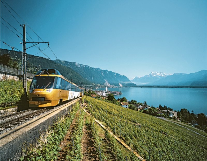 Imagem do trem da Golden Pass durante o dia com o trem do lado esquerdo da imagem e do lado direito gramado e ao fundo casas e um rio. Representa como viajar de trem pela Suíça.