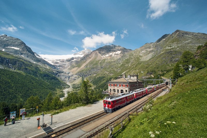 Imagem do trem Bernina durante o dia com a ferrovia do lado esquerdo com um trem em movimento e ao fundo montanhas. Representa como viajar de trem pela Suíça.