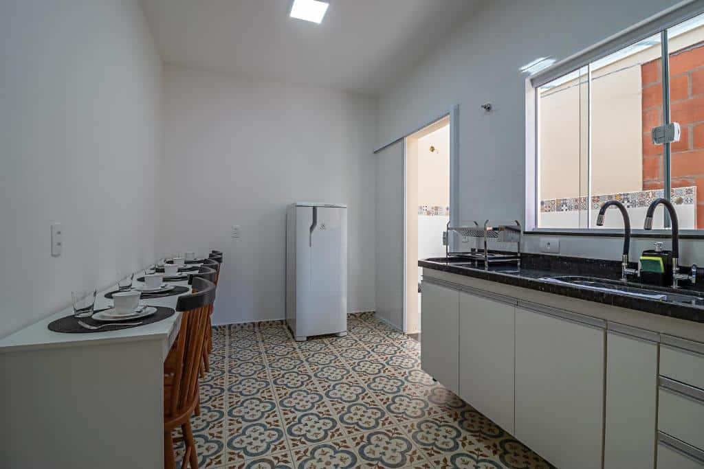 Cozinha do Vila Pepira ViP com uma mesa retangular comprida encostada na parede do lado esquerdo com mesas e utensílios em cima. Já do lado direito tem uma pia para lavar louça com uma janela de vidro. Já ao fundo tem uma geladeira com uma porta de vidro ao lado, imagem ilustrando post Airbnb em Brotas.