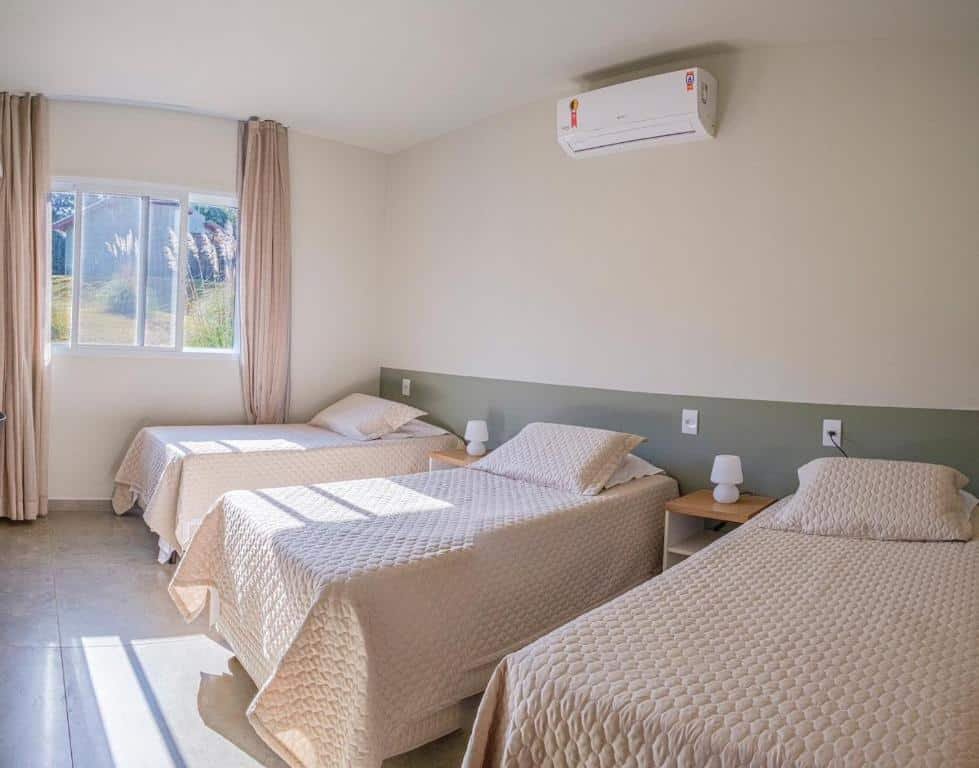 Quarto da casa do Vila Poção com três camas de solteiro, uma mesinha com um pequeno abajur do lado de cada cama e três tomadas. No quarto tem um ar-condicionado na parede e ao fundo uma janela de vidro que entra a claridade do dia. Imagem ilustrando post Airbnb em Brotas.