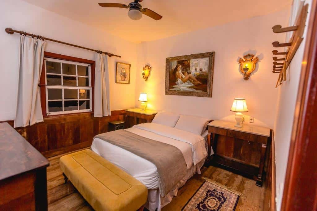 Um dos hotéis fazenda em Minas Gerais, a Vila Relicário. Na foto há uma cama box de casal no centro, e ladeando ela nos dois lados há uma mesa de cabeceira com abajur. Na parede atrás da cama há um quadro no centro da parede. Na parede à esquerda há uma janela com cortinas.