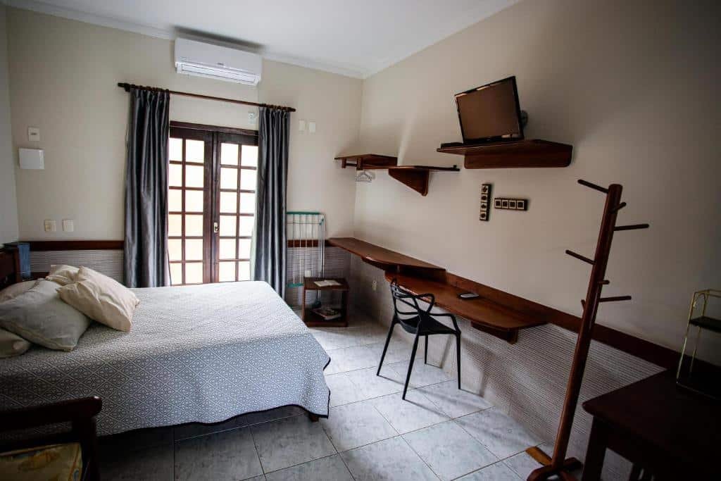 Quarto da Villa Bela Flats Ubatuba. Uma cama de casal do lado esquerdo, do lado direito um cabideiro, uma mesa, prateleiras e uma televisão. No fundo uma porta com cortinas. Foto para ilustrar post sobre airbnb na Praia da Enseada.