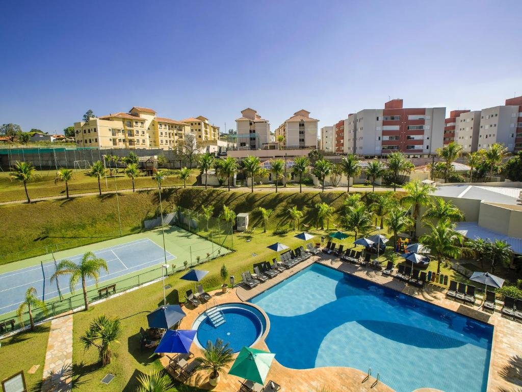 Piscina do Vinhedo Plaza Hotel com uma quadra de tênis ao lado, muitas árvores e cadeiras com guarda-sóis perto da piscina, para representar hotéis perto do Hopi Hari