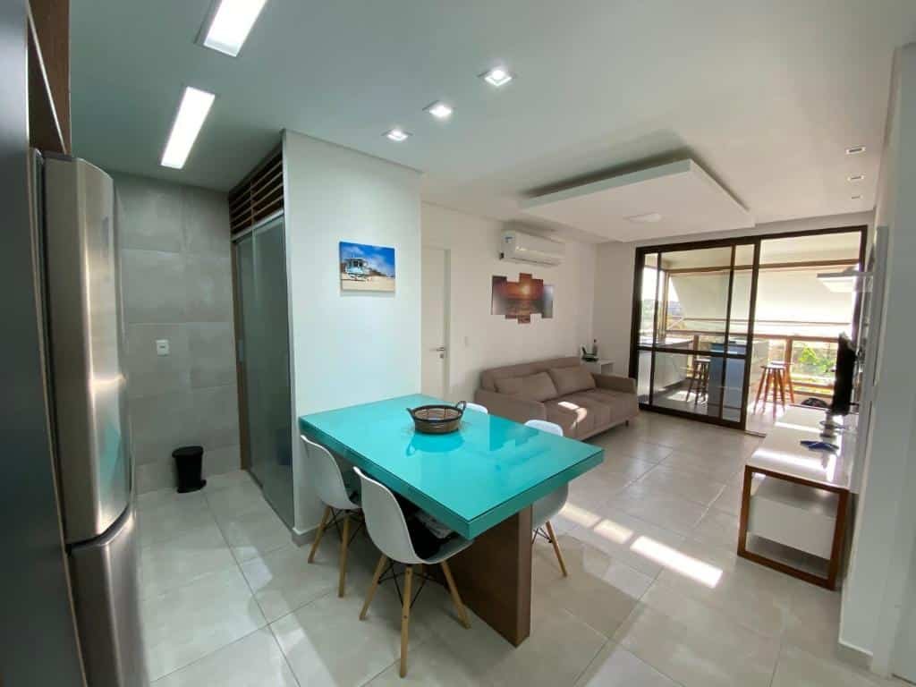 Sala e cozinha do Wai Wai Cumbuco Residence. Na sala há um sofá encostado na parede e de frente para uma tv. Ao fundo há uma porta levando para outro ambiente. Em primeiro está a cozinha com uma mesa de quatro lugares e uma geladeira de frente paar ela. Este é um dos airbnb em Fortaleza.