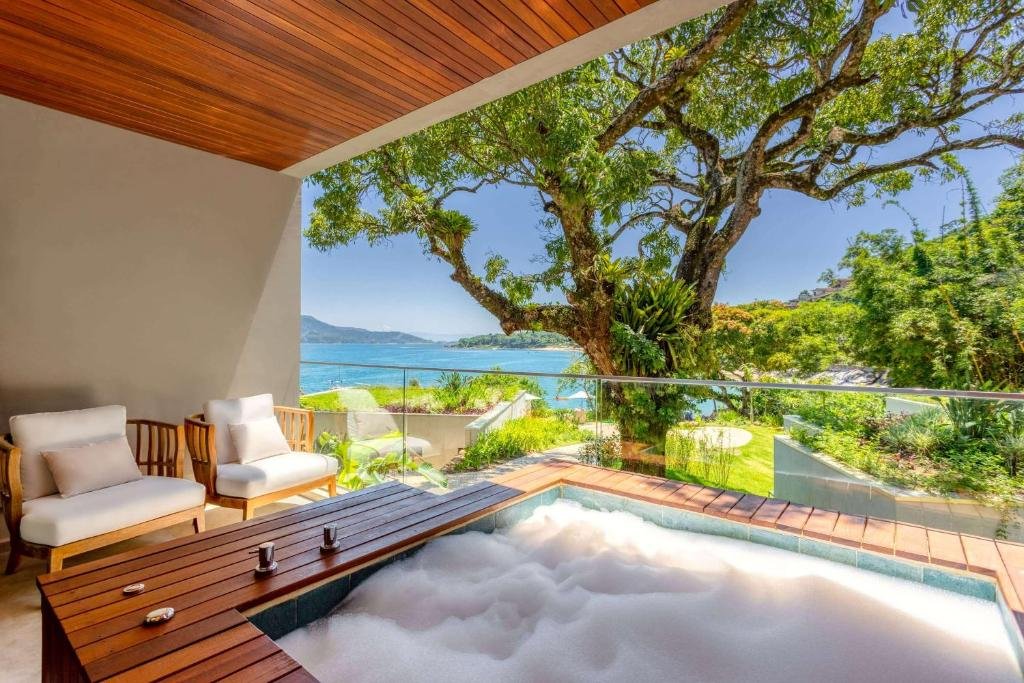 Varanda de um dos quartos do hotel Wyndham Ilhabela Casa Di Sirena. Na frente da imagem há uma banheira cheia de espuma e duas cadeiras no lado esquerdo da varanda. Já ao fundo a vista da varanda é para uma vasta vegetação a direita e para o mar a esquerda. Imagem utilizada para ilustrar o post pousadas pet friendly em Ilhabela.