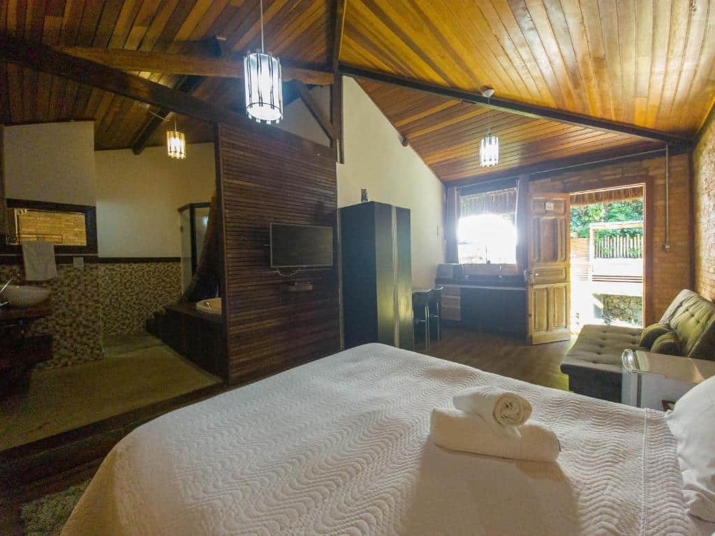 Foto de um quarto no Yannai Chalé Praia. A cama box e de casal está encostada na parede à direita. Do lado dela, mais ao fundo da foto, há um sofá. E mais ao fundo da foto há a porta de entrada e uma cozinha compacta. De frente com o sofá há um painel de TV. Na esquerda está o banheiro com hidro.