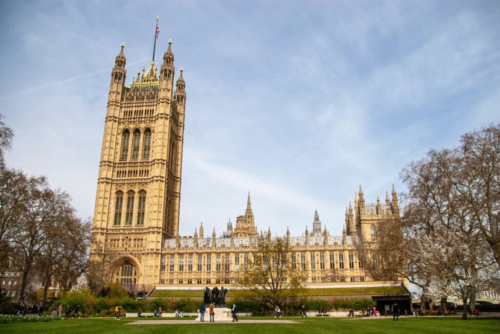 A imagem mostra o Palácio de Westminster, em Londres, em um dia claro, com visitantes no gramado e nas árvores ao redor da área. Essa é uma das opções de o que fazer em Londres.