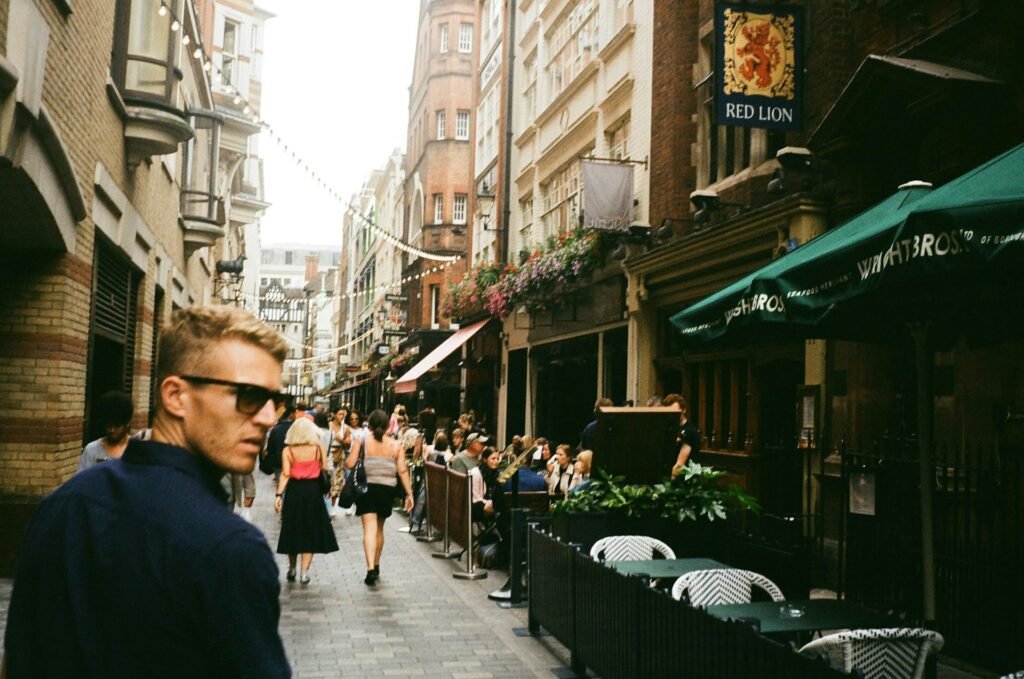 Uma movimentada rua europeia com restaurantes ao ar livre, luzes penduradas e um homem de óculos escuros caminhando, perto de um pub com placa "Red Lion". Esse é o Soho, um dos bairros recomendados no roteiro para Londres.