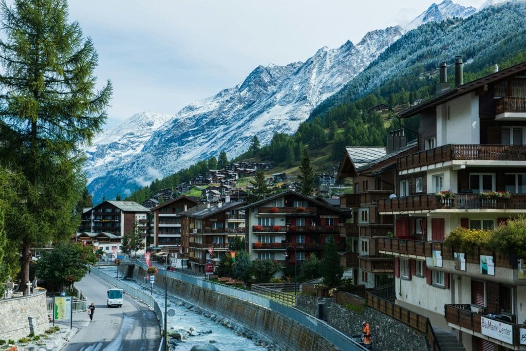 Imagem da cidade do Zermatt, Suíça durante o dia com um rio no centro e do lado direito vários prédios e ao fundo atrás dos prédios montanhas. Representa o que fazer na Suíça.