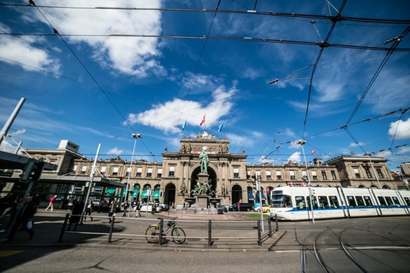 Imagem durante o dia da Estação central em Bahnhofstrasse com um edifício ao fundo e em frente dele uma avenida e do lado direito da imagem um trem. Representa o que fazer em Zurique.