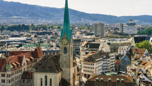 Vista aérea de Zurique com uma torre de igreja proeminente, cercada por edifícios históricos e modernos, com colinas ao fundo.